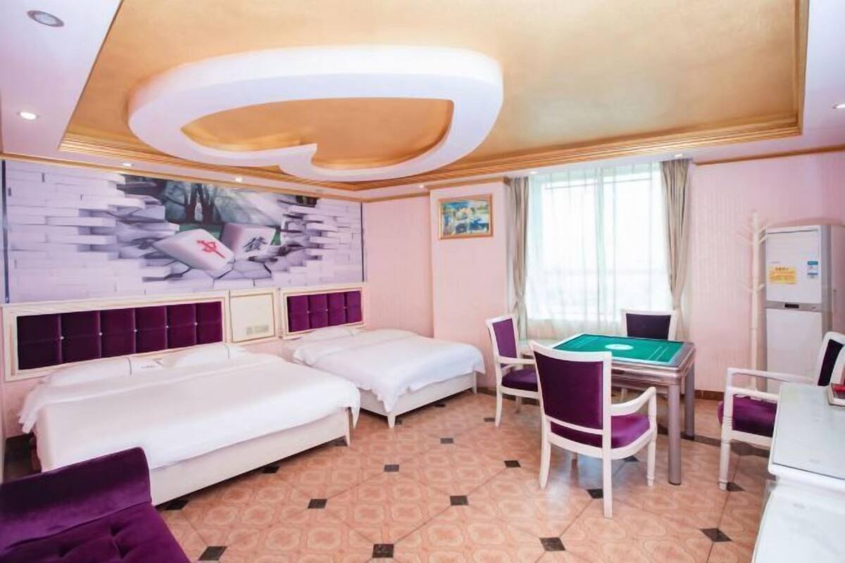 A hotel room with two beds, a dining table with chairs, a wall-mounted TV, and a ceiling light fixture.
