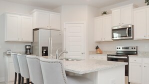 A modern kitchen with white cabinets, a stainless steel refrigerator, a microwave, and an oven. There are three bar stools at the island. - Backwater Bliss (Port St. Joe)