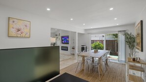 Un espace repas moderne avec une table et des chaises en bois, un téléviseur fixé au mur et une vue sur le jardin grâce à des portes vitrées.