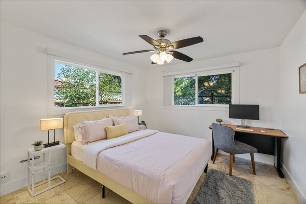 A bedroom with a bed, desk, chair, ceiling fan, and windows.
