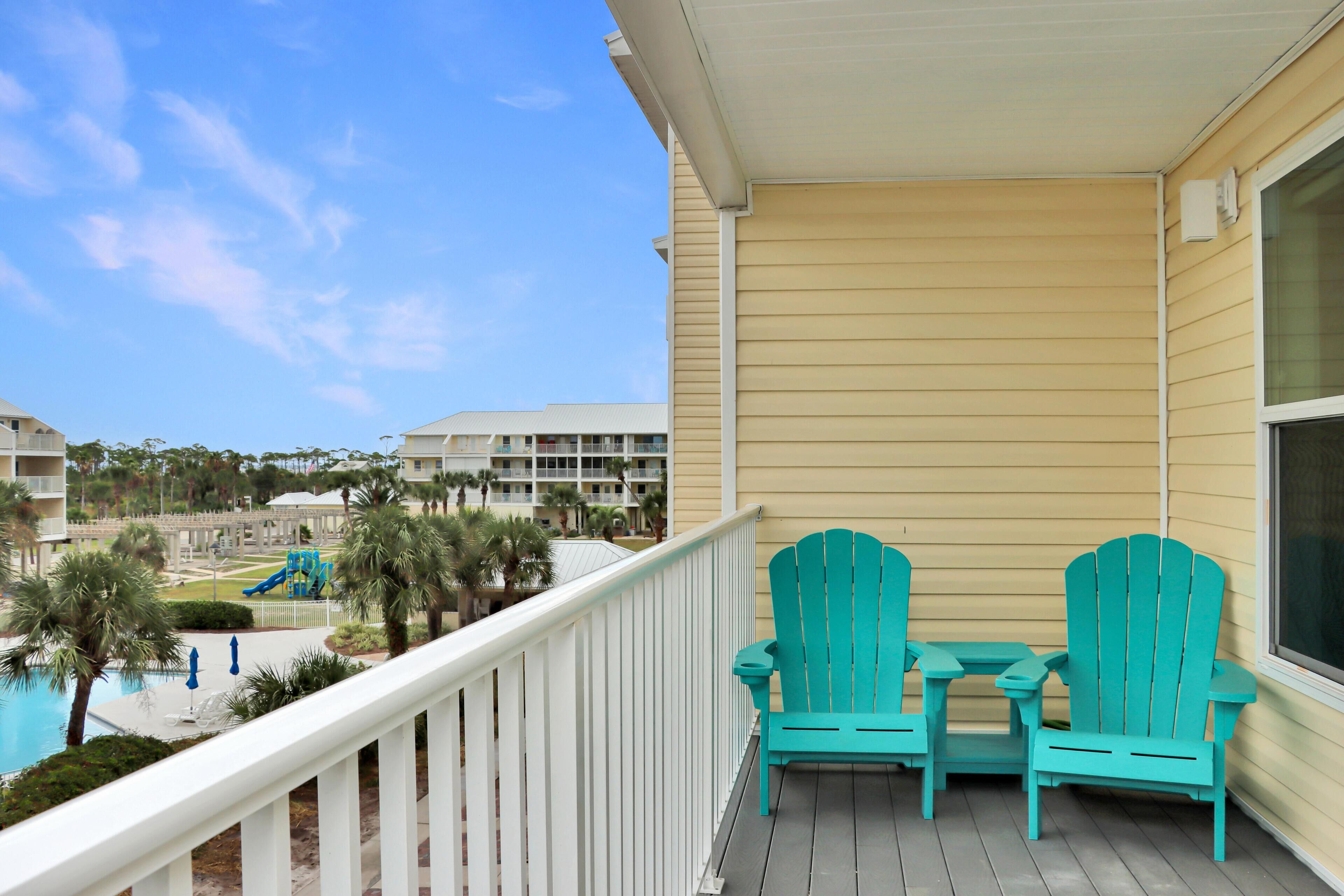 A balcony with two turquoise Adirondack chairs and a small table, overlooking a pool and a playground area with palm trees.