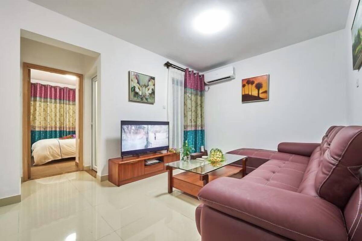A living room with a sofa, a TV, a coffee table, and a bed in the background.