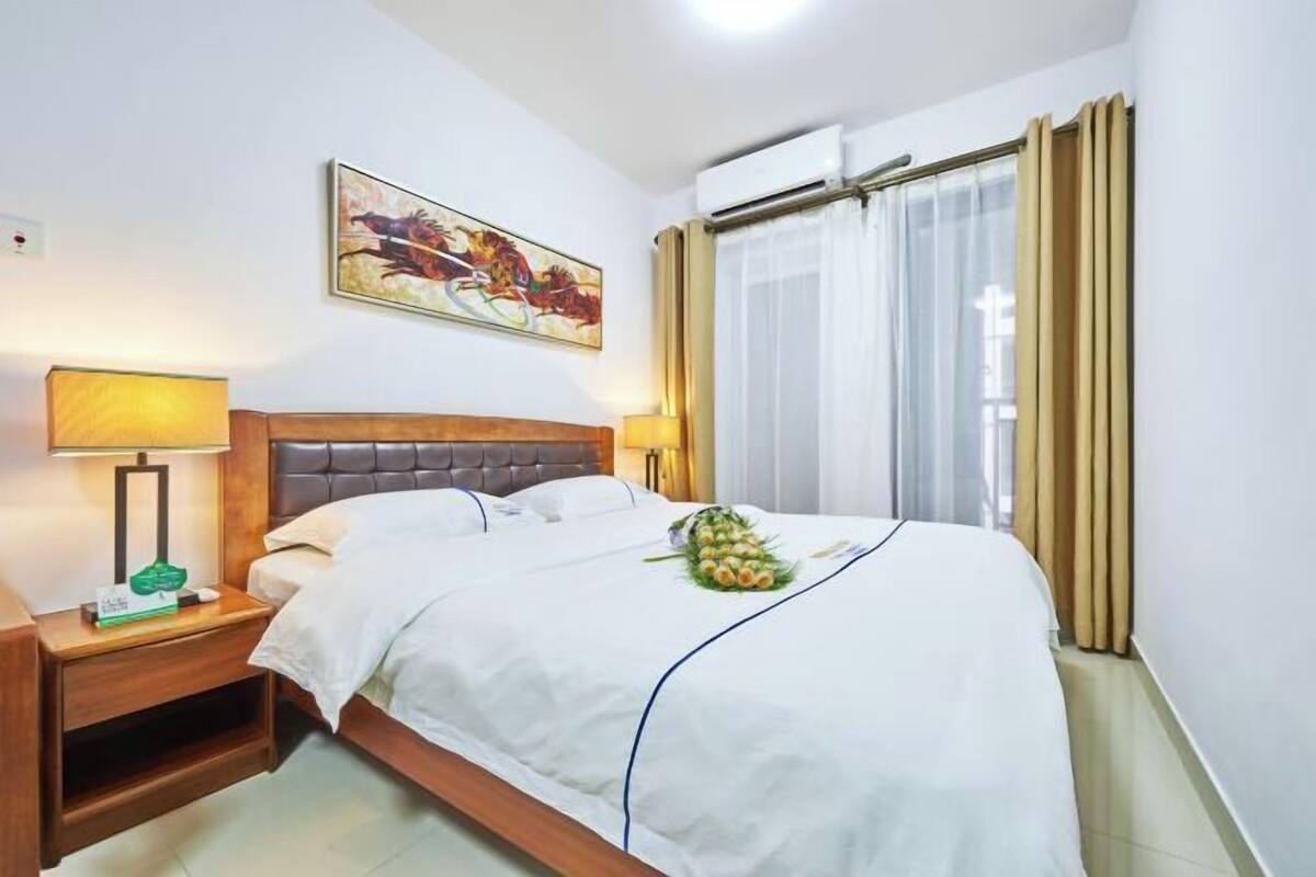 A hotel room with a bed, two bedside tables, a wall-mounted air conditioner, a window with curtains, and a painting on the wall.
