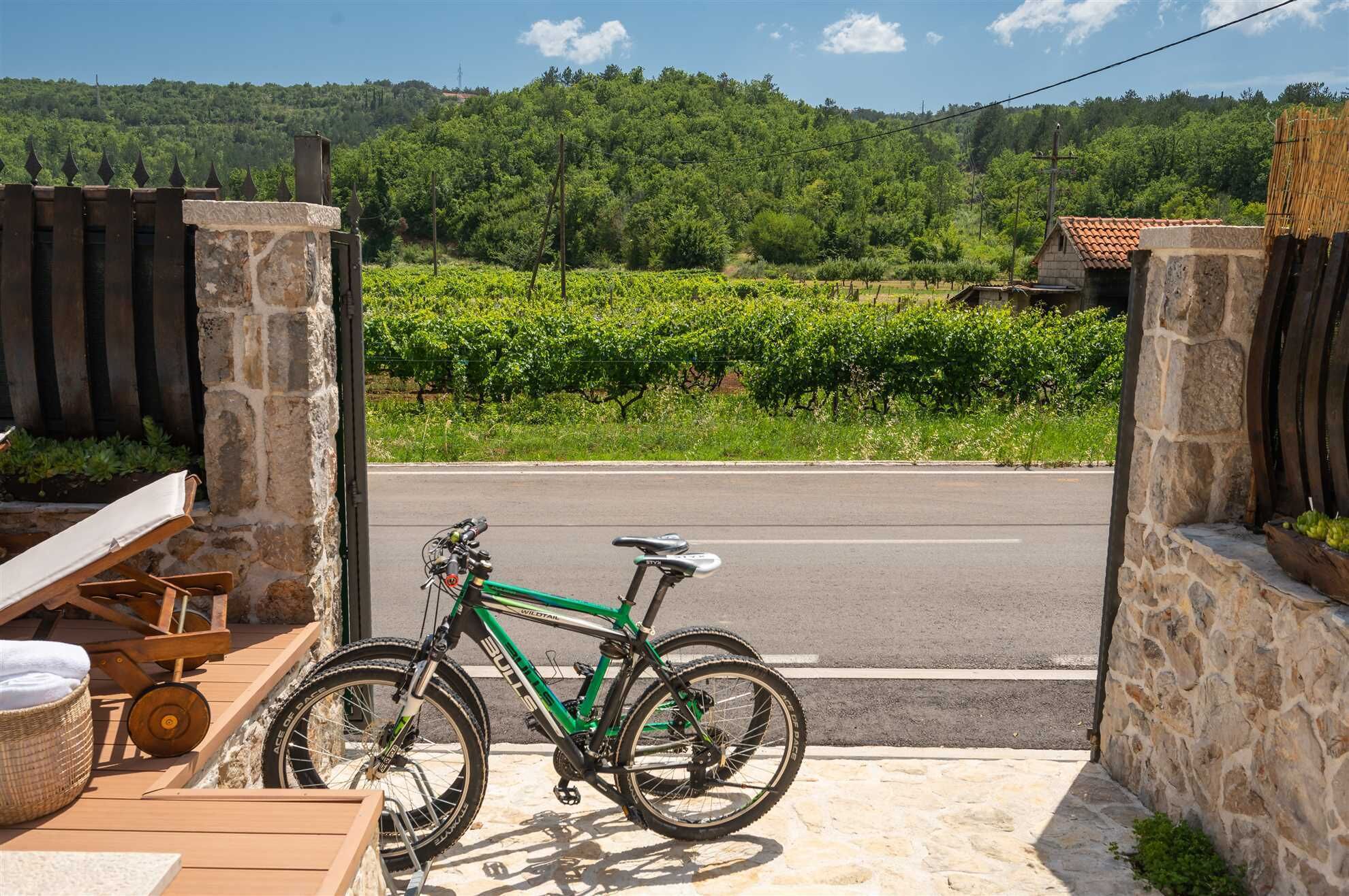 A bicycle parked in a stone-walled outdoor area with a view of a road and vineyards in the background.