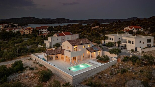 Une villa moderne dotée d’une piscine, entourée d’un paysage vallonné et bordée, au loin, par une étendue d’eau.