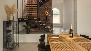 Un espace repas moderne avec une table en bois, des chaises noires et un lustre noir. Un escalier avec un mur en briques et une rampe en métal mène à un niveau supérieur.