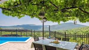 A poolside area with a table and chairs, a view of mountains, and a pergola with hanging lights.