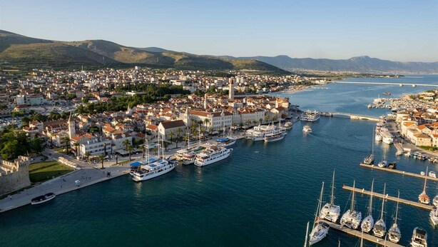 An aerial view of a coastal town with a marina, buildings, and hills in the background.
