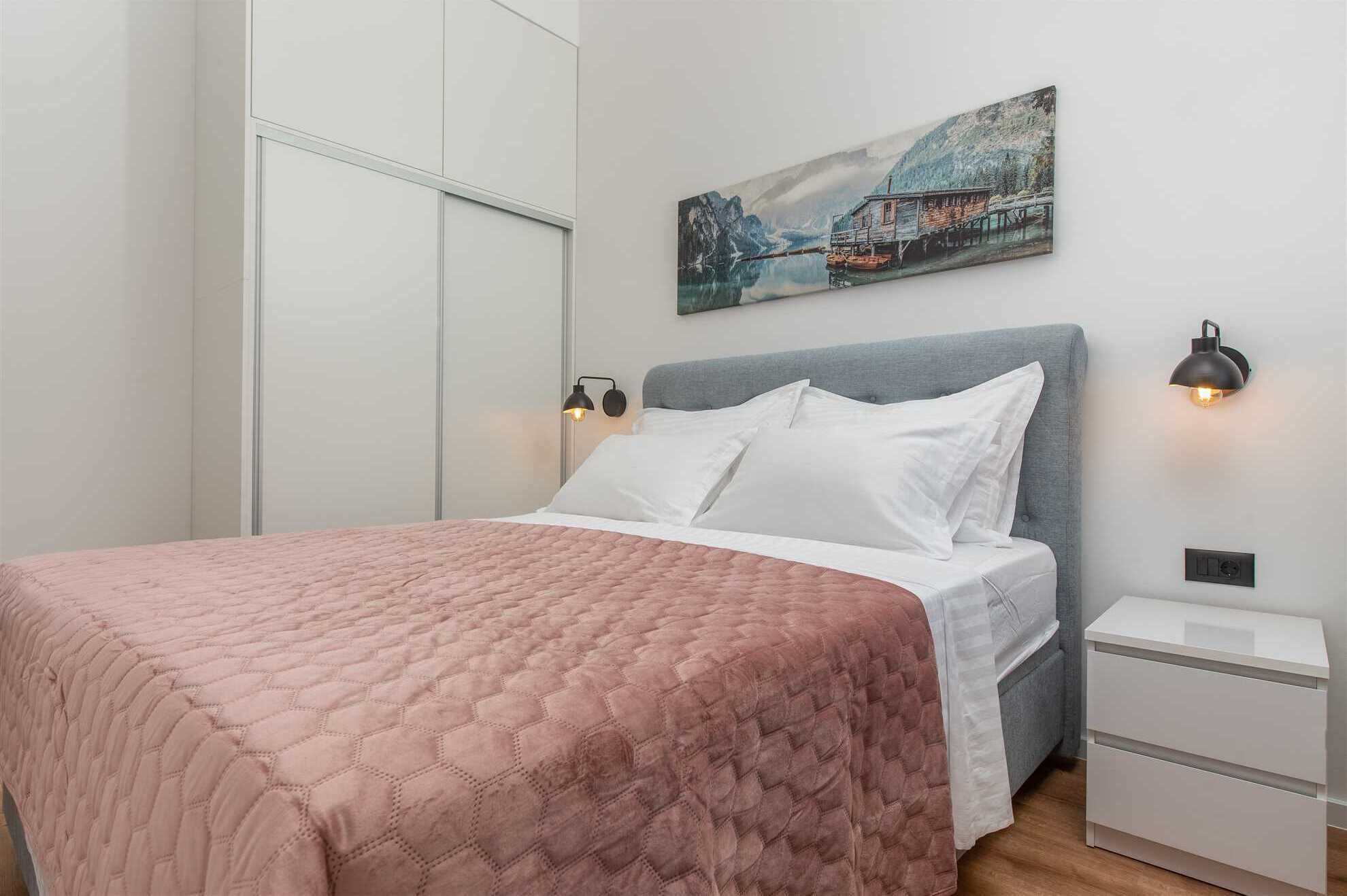 A neatly made bed with a pink quilt, a grey headboard, and a painting of a lakeside scene above it.