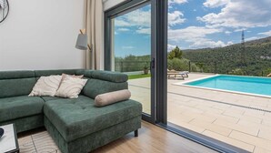 A modern living room with a green sofa, a large sliding glass door leading to a pool area with a view of a hilly landscape. - Villa Nacle with heated Pool near Omiš (Tugare)