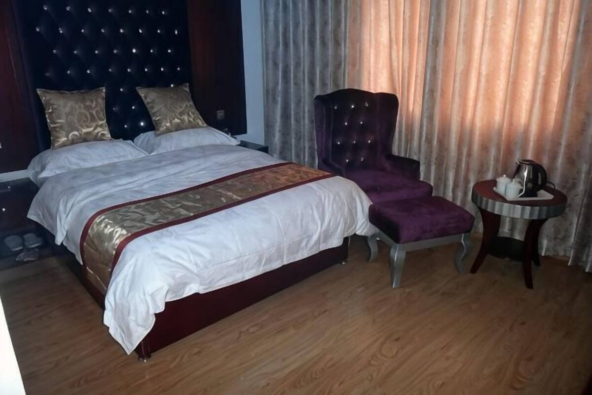 A double bed with a headboard, a chair, a small round table with a kettle, and curtains.