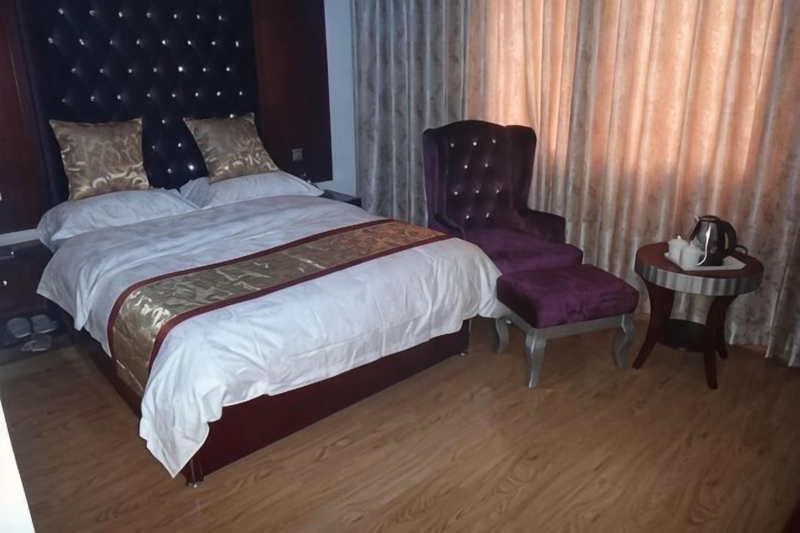 A double bed with a headboard, a footstool, a small round table with a kettle, and a curtain backdrop.
