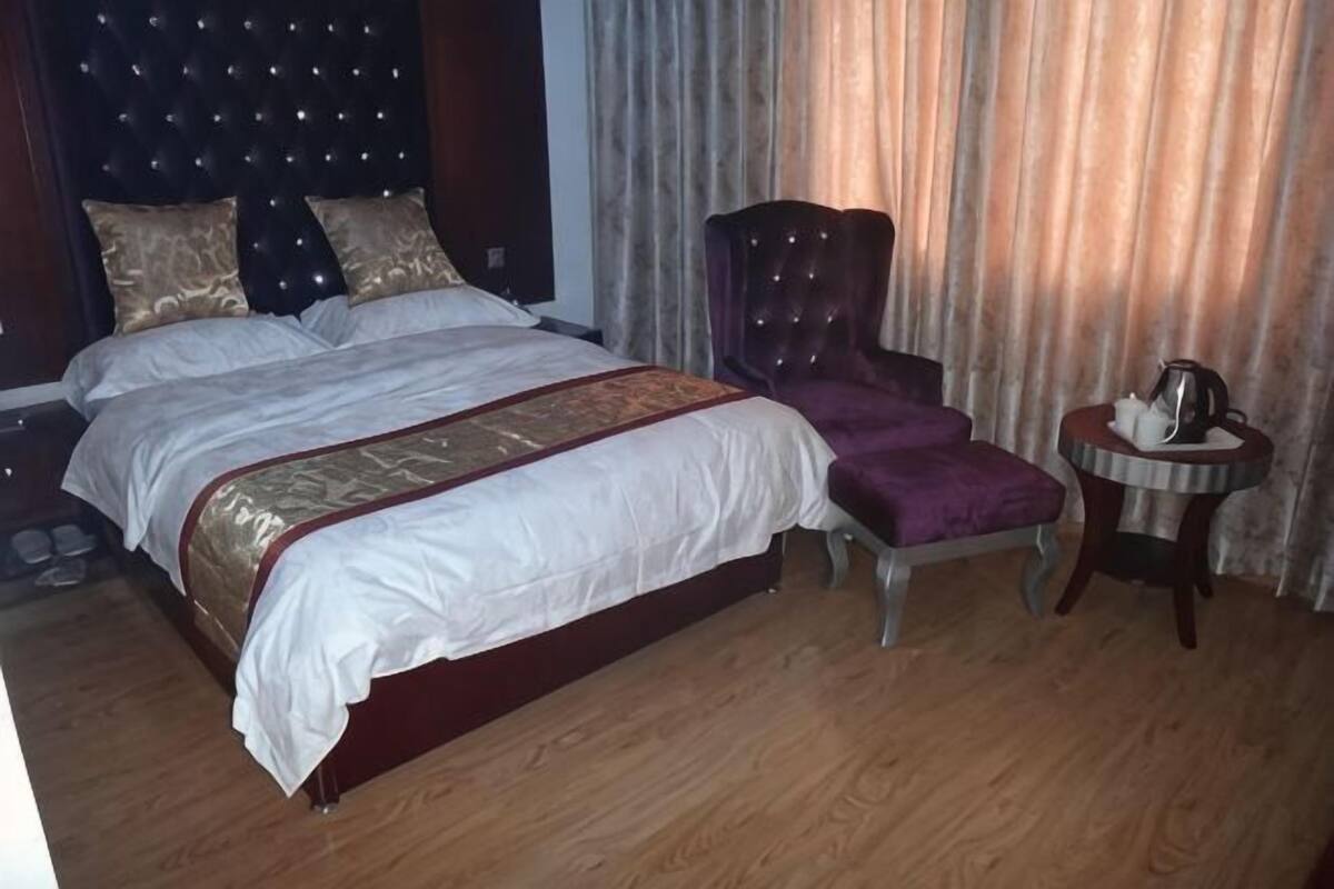 A double bed with a headboard, a footstool, a small round table with a kettle, and a curtain backdrop.