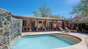 A swimming pool with a stone wall on one side, a covered outdoor seating area, and a building with multiple doors and windows.