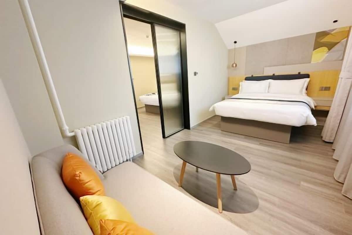 Une chambre d’hôtel moderne, équipée d’un lit, d’un canapé avec des coussins orange, d’une petite table ronde et d’une porte coulissante donnant sur une autre pièce.