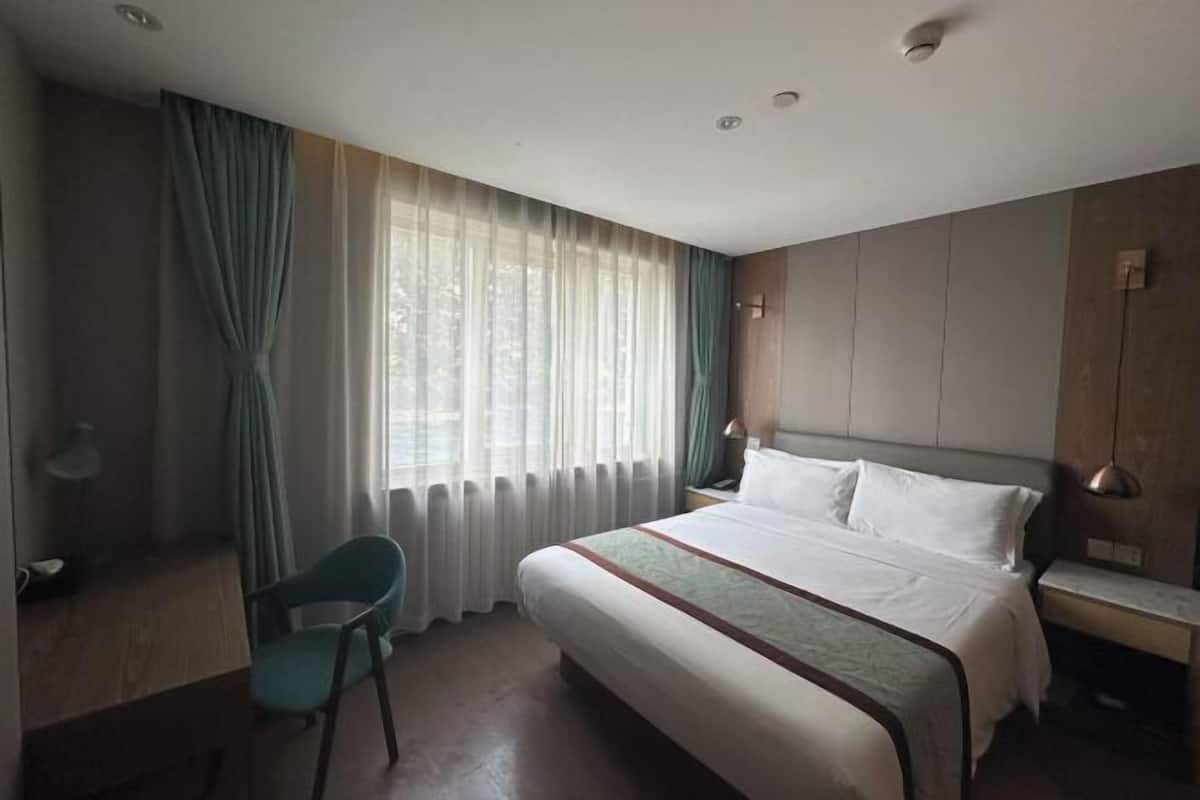 Une chambre d’hôtel avec un lit, une chaise, un bureau, une lampe, une table de chevet et une fenêtre avec des rideaux.