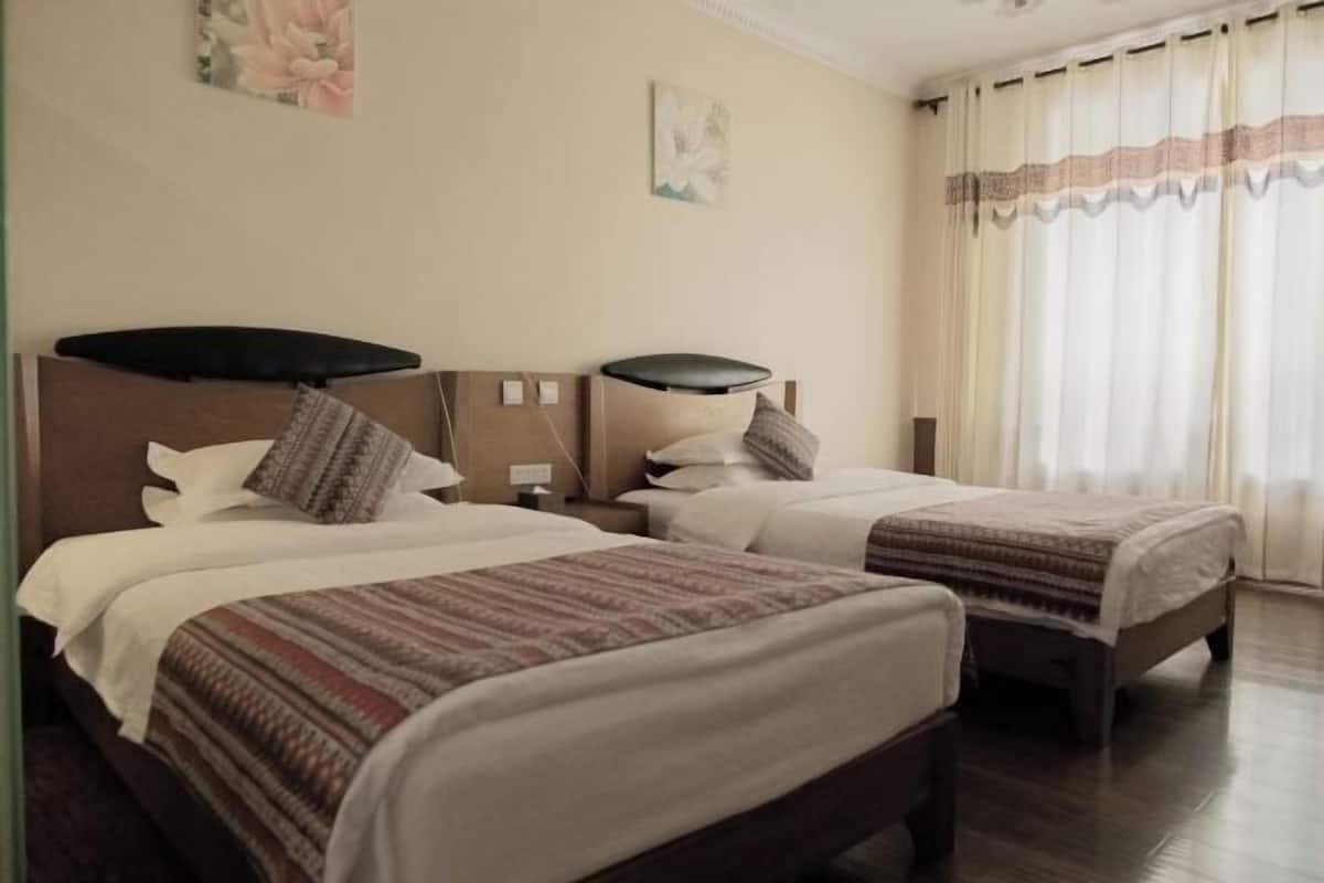 A hotel room with two beds, a wooden headboard, a nightstand, a lamp, and a window with curtains.