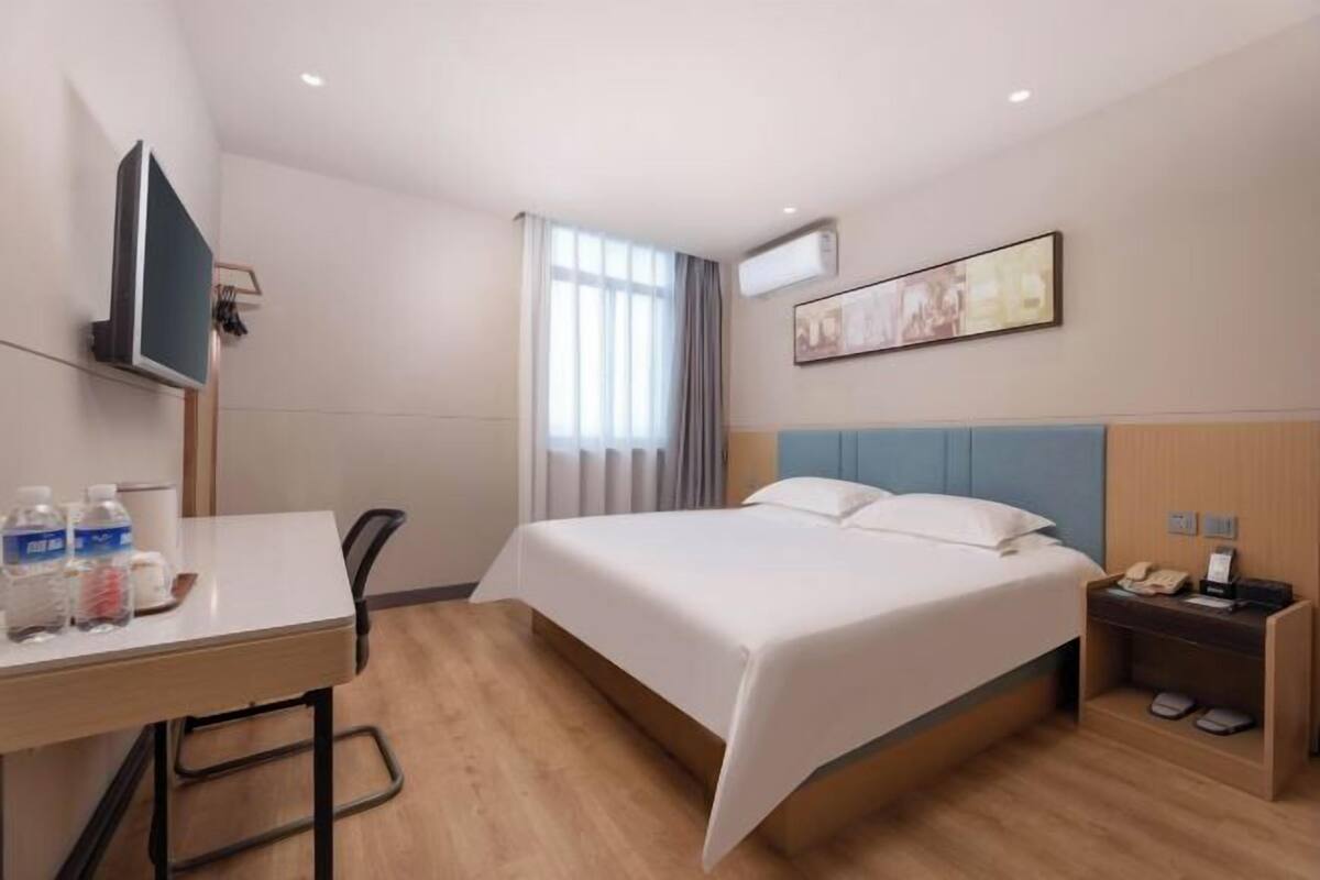 Ein Hotelzimmer mit einem Bett, einem Schreibtisch mit Stuhl, einem Fernseher, einem Fenster mit Vorhängen und einer Wand-Klimaanlage.