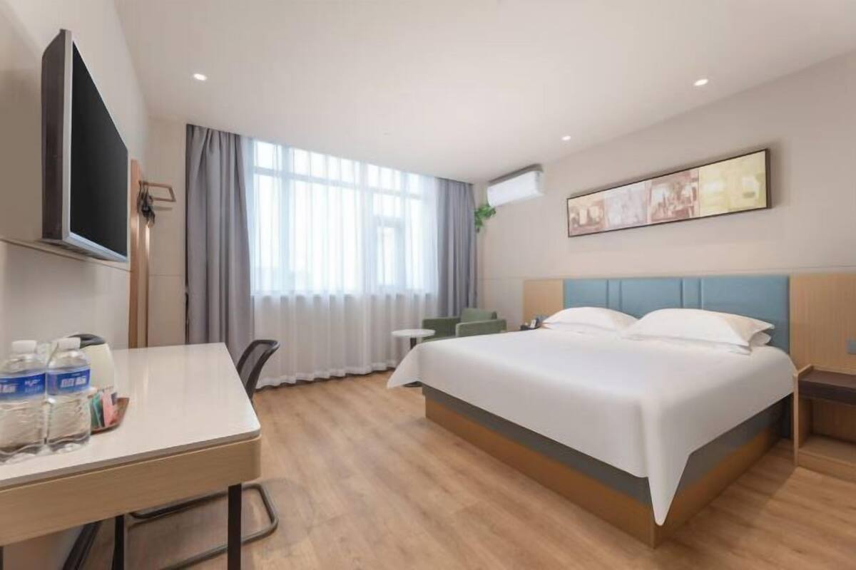 Ein Hotelzimmer mit einem Bett, einem Schreibtisch mit Wasserflaschen, einem Fernseher, einem Fenster mit Vorhängen und einem Bild an der Wand.
