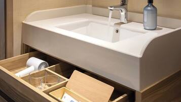 A bathroom with a white sink, a faucet, a soap dispenser, and a wooden drawer with a hairdryer and a glass.