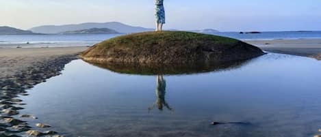 Une personne se tient debout sur une petite île, les bras levés, entourée d’une eau calme et sous un ciel dégagé.