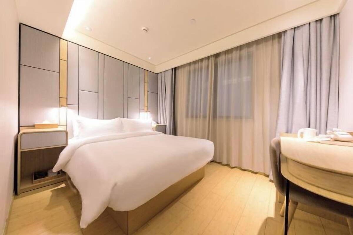 Habitación de hotel con cama, mesita de noche, lámpara y vista al exterior a través de cortinas.