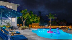 Un área de piscina con sillones reclinables, una terraza de madera y un cielo nocturno.