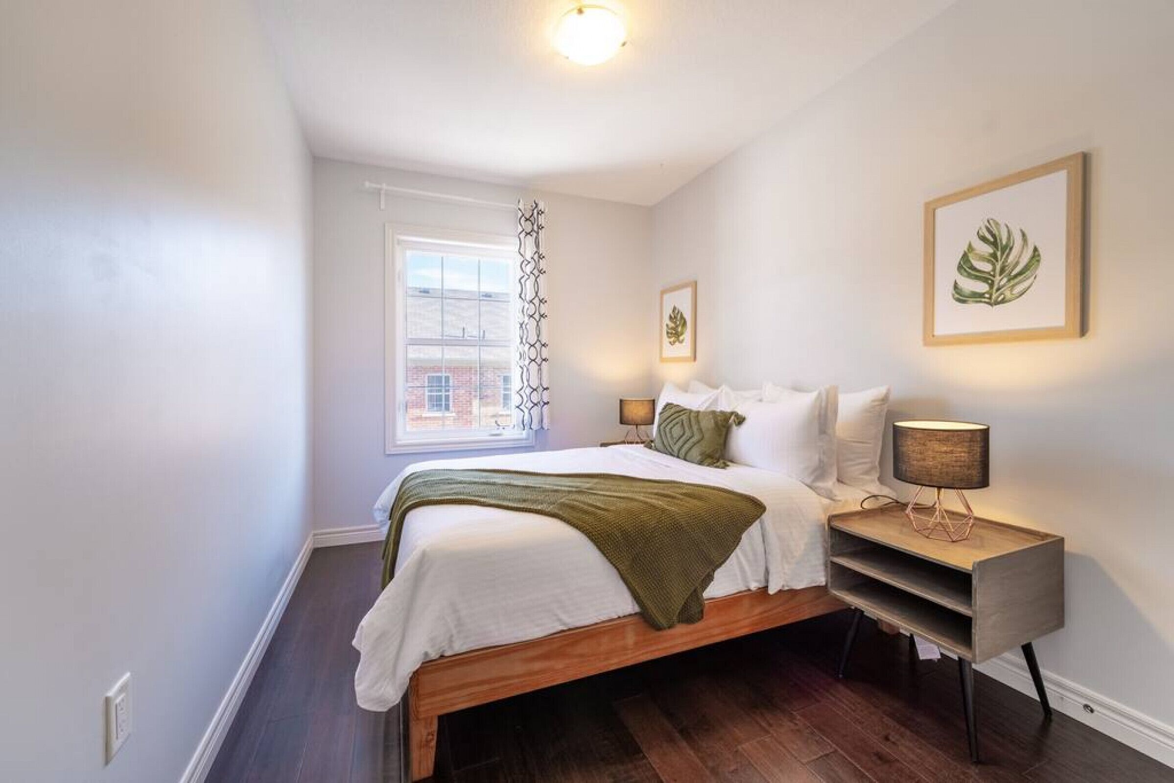A bedroom with a bed, bedside lamps, a window, and a framed plant.