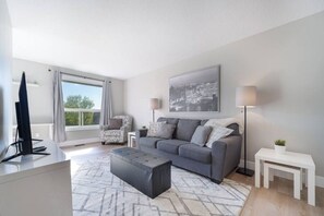 A modern living room with a grey sofa, a coffee table, a TV, and a large window. - Bright & Spacious - 3-Bedroom Home (Kitchener)