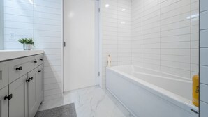 A bathroom with white tiled walls, a white bathtub, and a white vanity with drawers. - Bright & Cozy 1BR Basement Unit (Kitchener)