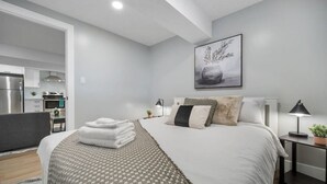 A modern bedroom with a neatly made bed, bedside lamps, and a kitchen visible in the background. - Bright & Cozy 1BR Basement Unit (Kitchener)
