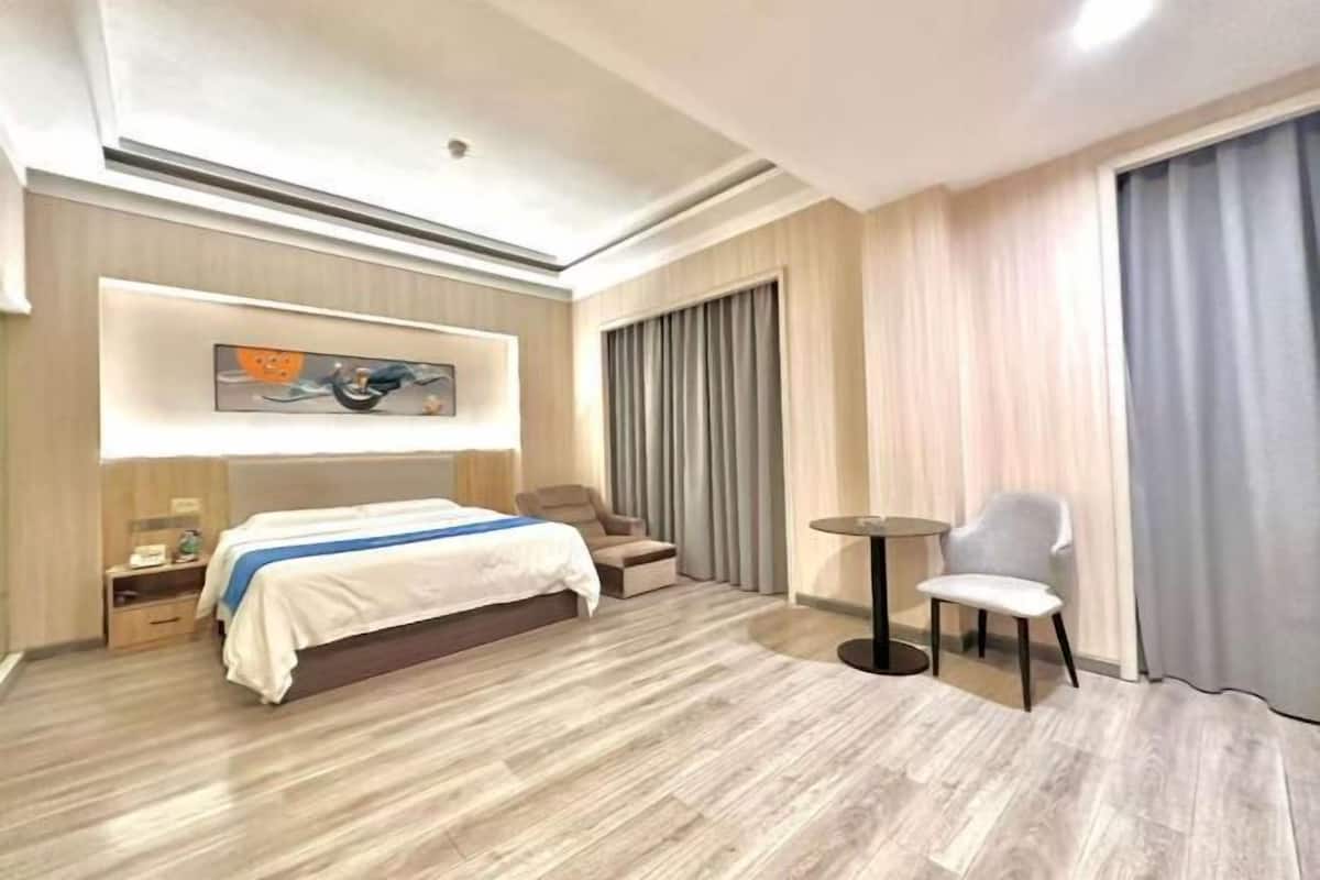 A hotel room with a bed, a chair, a small table, a painting on the wall, and a window with curtains.