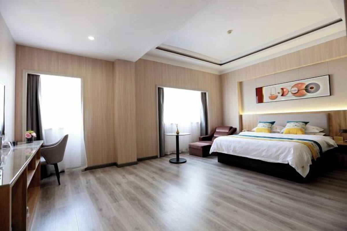 A hotel room with a bed, a desk, and a chair. There is a window with curtains, a wall-mounted artwork, and a ceiling light.