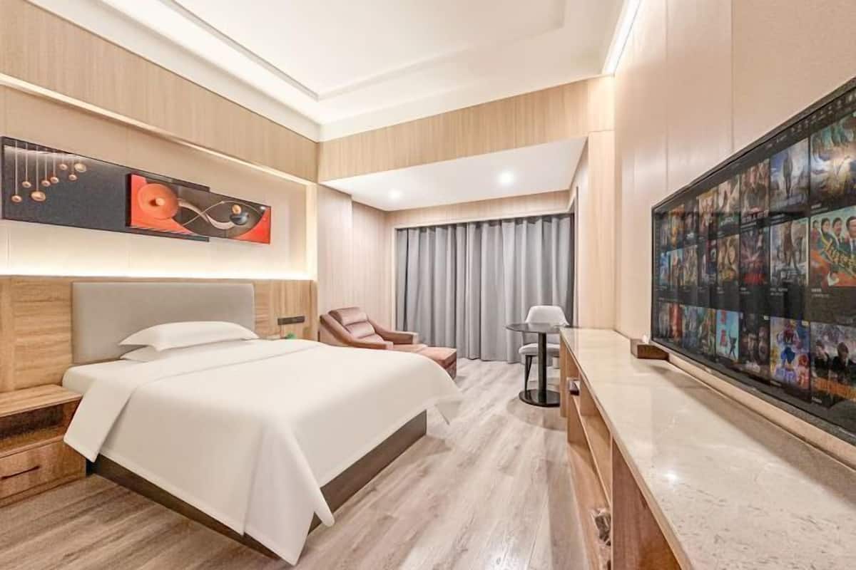 A modern hotel room with a large bed, a seating area with a chair, a small table, and a wall-mounted TV displaying movie posters.