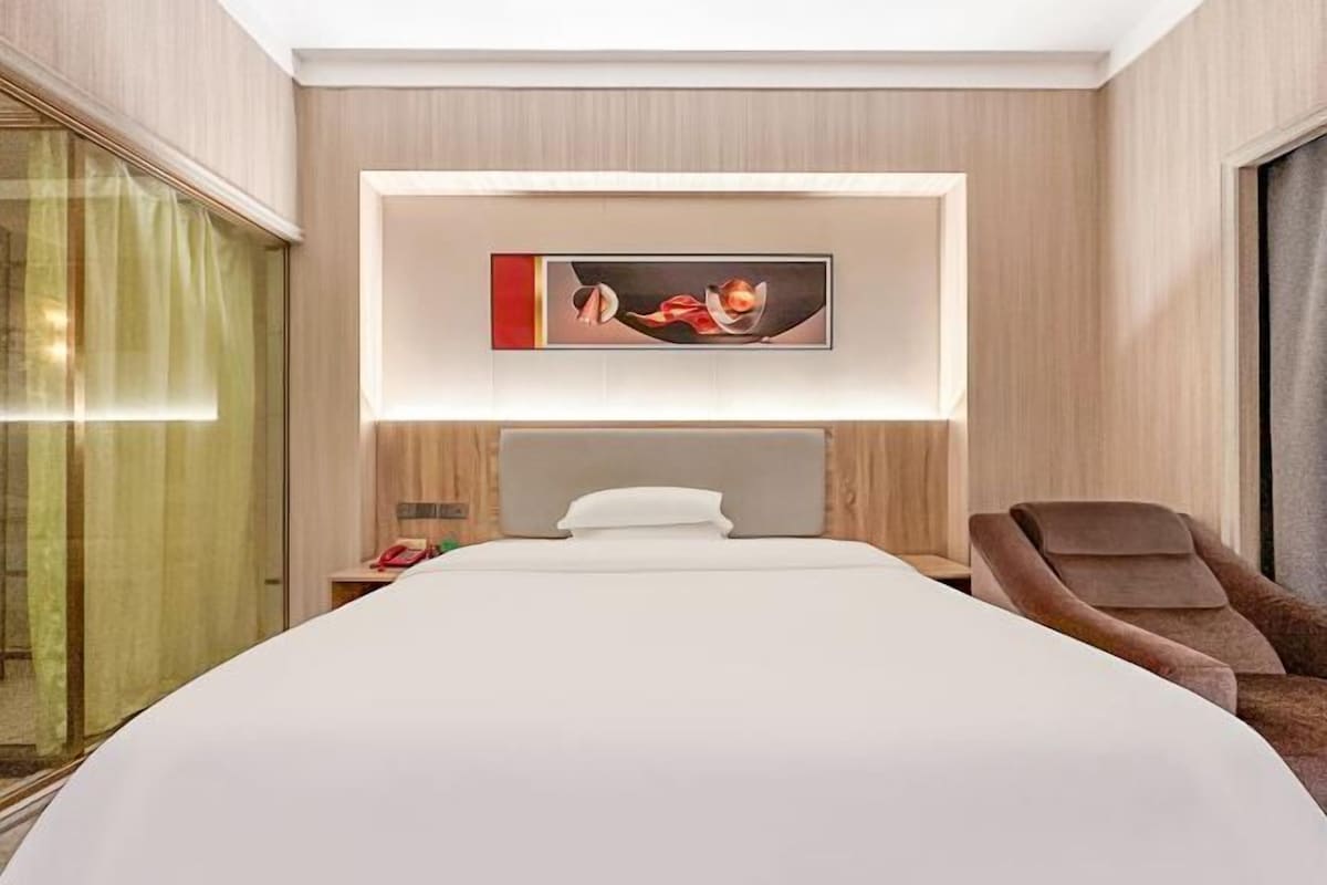 A modern hotel room with a large bed, a chair, a bedside table, and a wall-mounted artwork.