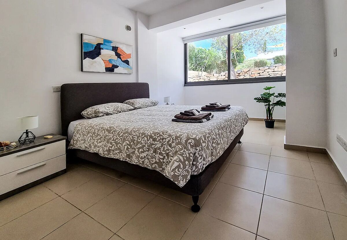 A bedroom with a large bed, a nightstand, a lamp, a window with a view of trees, and a painting on the wall.