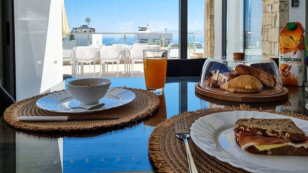Et bord med morgenmadsprodukter, blandt andet brød, en skål og et glas appelsinjuice, med udsigt til havet og bygninger i baggrunden.