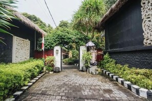 Un camino pavimentado conduce a una entrada con portón, con una estructura de techo de paja a la izquierda y una pared negra con detalles decorativos a la derecha.