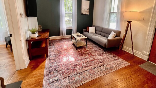 Un salon avec un mur vert foncé, un canapé gris, une table basse en bois, un tapis rouge et beige, et un lampadaire.