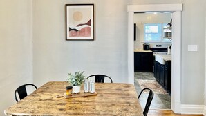 Un coin repas avec une table en bois rustique, des chaises, une œuvre d’art encadrée, et une cuisine moderne en arrière-plan.