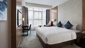 Habitación de hotel con una cama grande, un escritorio, una silla y vistas de la ciudad a través de amplios ventanales.