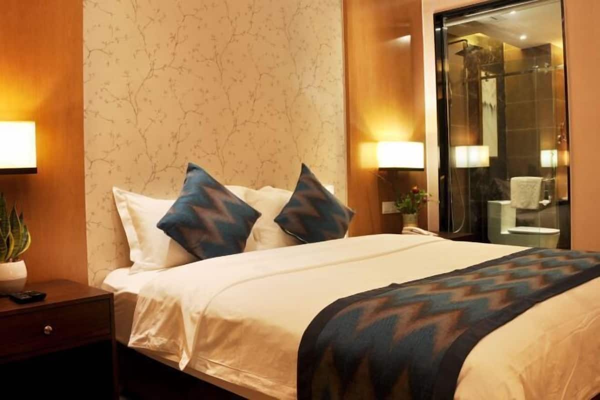 A hotel room with a bed, two pillows, a nightstand with a plant, and a bathroom visible through a glass door.