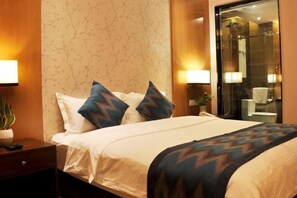 Ein Hotelzimmer mit einem Bett, zwei Kissen, einem Nachttisch mit Pflanze und einem durch eine Glastür einsehbaren Badezimmer.