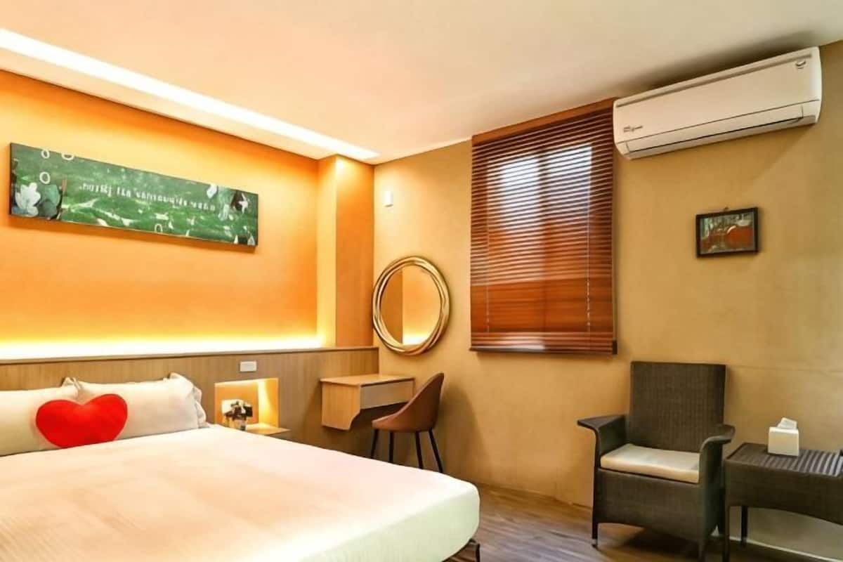 A hotel room with a bed, a desk, a chair, a round mirror, an air conditioner, and a window with blinds.