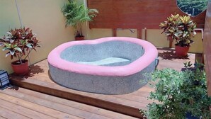 Ein runder Stein-Whirlpool mit rosa Rand, umgeben von Holzterrasse, Topfpflanzen und einem Holzzaun.