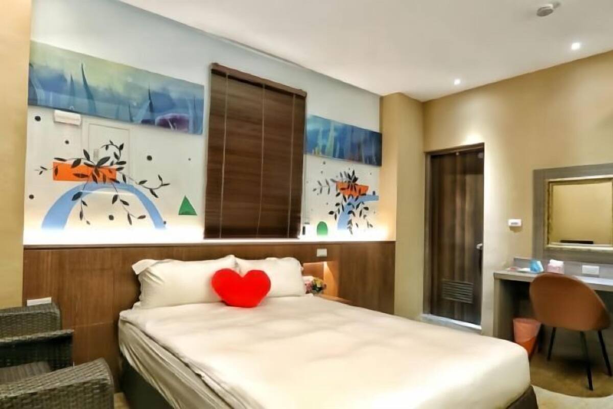 A hotel room with a bed, a desk, and a chair. There are two windows with blinds, a wall with artwork, and a door.