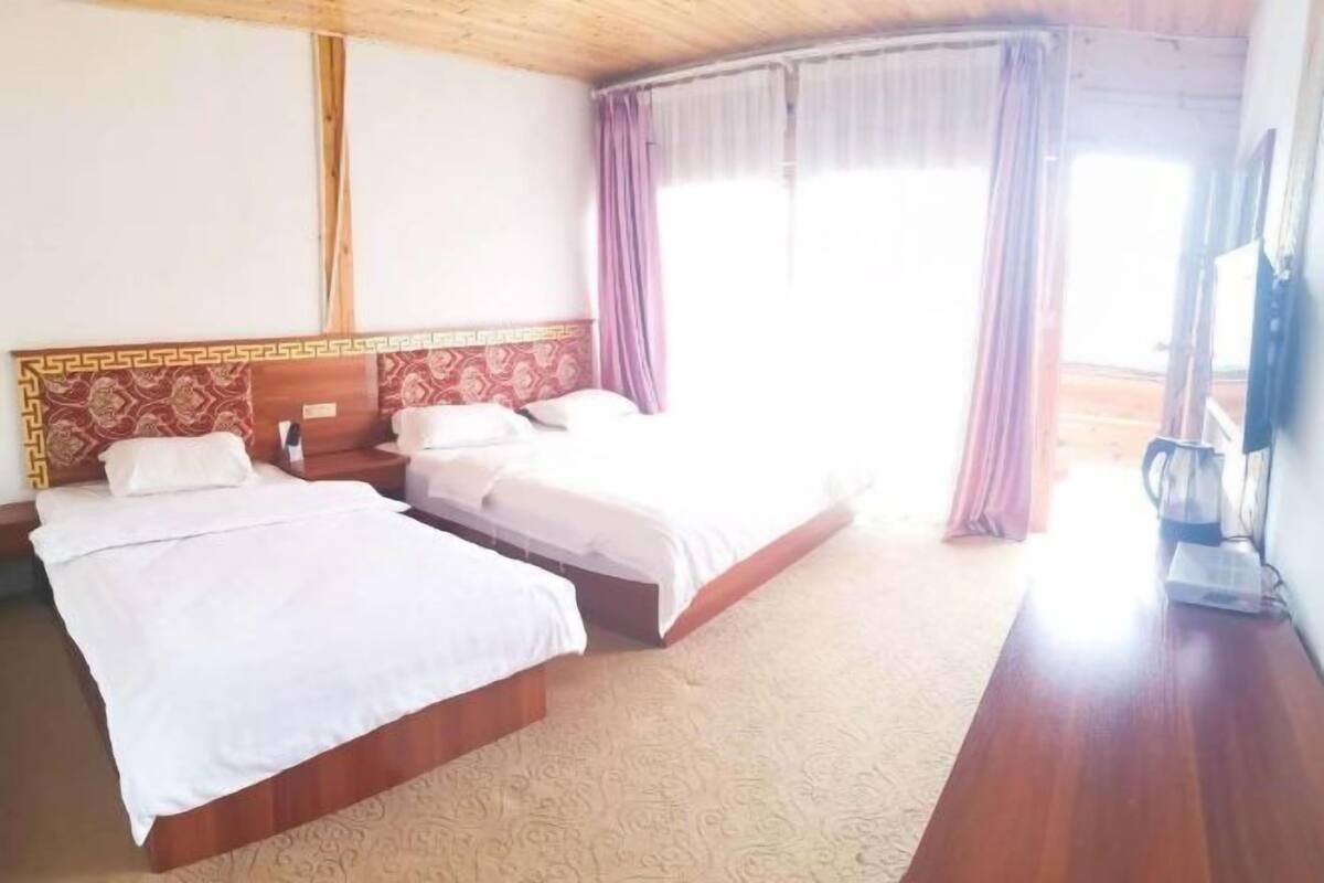 Ein Hotelzimmer mit zwei Betten, einem Holzkopfende, einem großen Fenster mit Vorhängen und einem Flachbildfernseher.