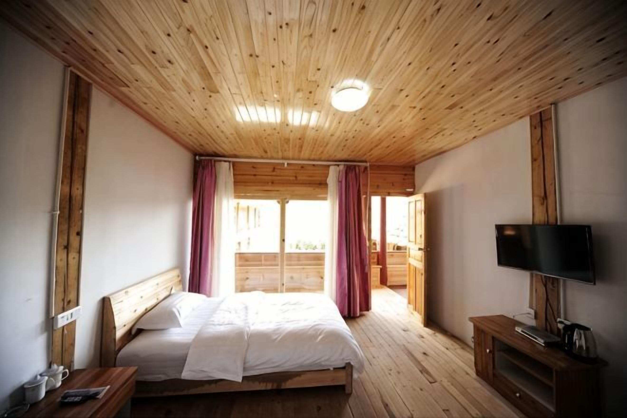 A bedroom with a wooden ceiling, a bed, a television, and a window with curtains.