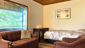 A cozy room with a wooden ceiling, a large window with a view of trees, a bed with a floral bedspread, and a brown sofa. - Small Cabin with gorgeous view and lake access (Nelson)