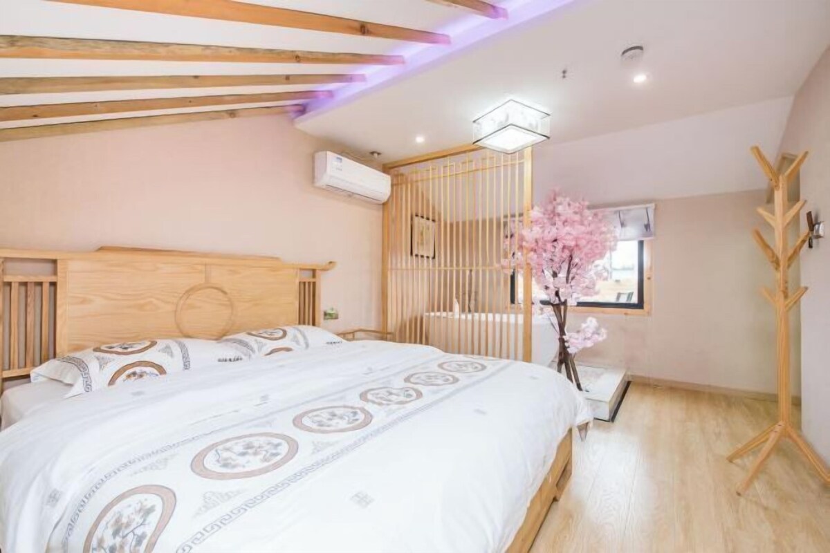 Quarto com cabeceira de madeira, cama com roupa de cama estampada, piso de madeira, um ventilador e uma janela com vista.